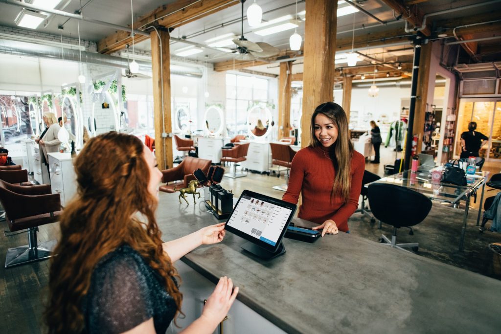 A lady taking payment from another lady within a beauty salon. The cashier is using a tablet POS system. There are various beauty stations in the background, with mirrors and chairs.