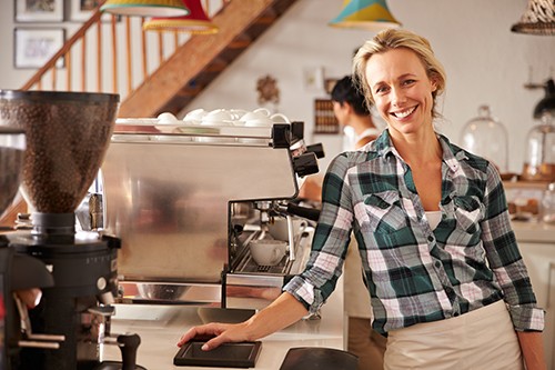 A blond, female barista smiling at the camera, standing beside a coffee machine and coffee grinder. She's wearing a checked shirt and a cream apron.