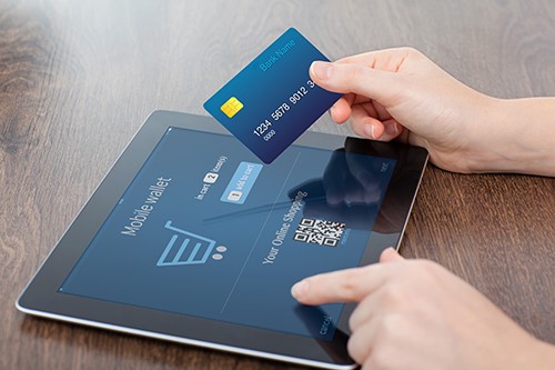 Two hands doing an online payment. The left hand is typing on a tablet, and the right hand is holding up a blue bank card.