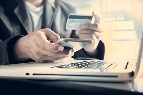 Somebody inputting their card details into their phone. The person is looking at their bank card, whilst typing on their phone and there is a laptop underneath their hands.