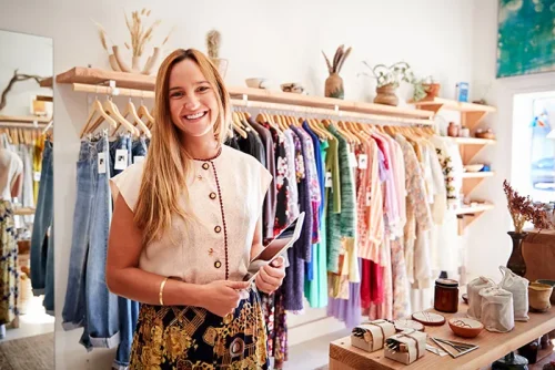 A blond lady who works in a fashion retail store is smiling at the camera, holding a tablet that is used to take payments. There are clothes hung up on hangers in the background.