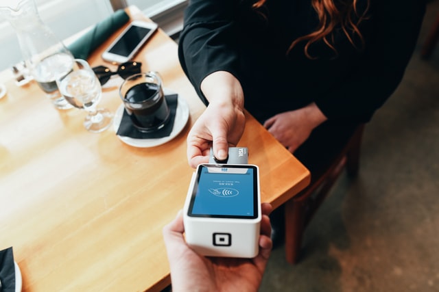 Taking a card payment using a card machine device. A lady is putting her card into the bottom of the machine. There is a coffee table in the background with various drinks on it.
