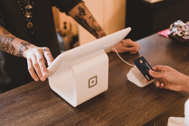 Taking a card payment at a till using a dedicated POS system. The cashier is wearing a black t shirt and has many tattoos. The customer is placing their debit card on the contactless part of the machine.