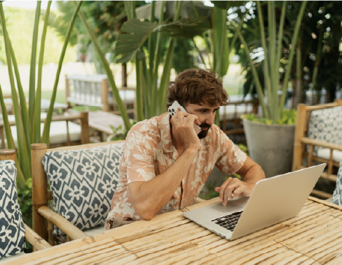A young man on the phone looking at his laptop in a remote looking environment.