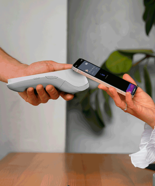 A contactless payment is taking place from a phone to a portable card reader machine. We can see a hand holding the machine and another holding the phone on the machine in order to make payment.