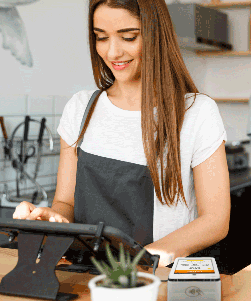A smiling lady is typing on a tablet, she’s wearing a white t-shirt and grey apron and there is a portable card reader machine to her left.