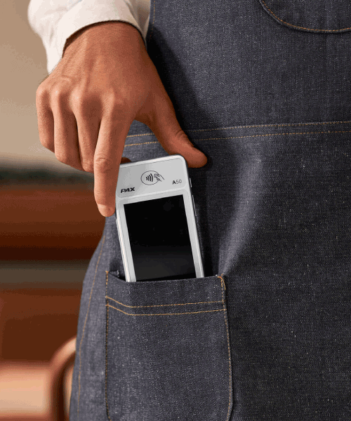 Someone is putting a portable card reader machine in the pocket of their denim apron.