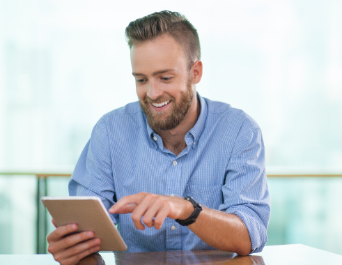 A young man with a beard smiling at a tablet he’s holding in his right hand. He’s wearing a blue checked shirt and a black watch on his left hand, which is pointing at the tablet screen.